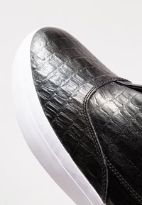 Gros plan sur une sneaker en cuir noir avec une texture crocodile et une semelle en caoutchouc blanc, sur fond blanc uni.