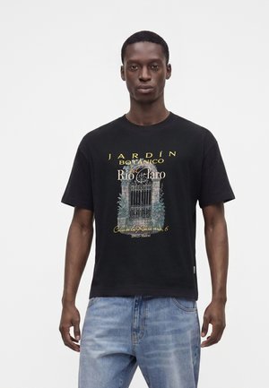 Jeune homme portant un t-shirt noir avec un graphique de portail de jardin et un texte en espagnol, associé à un jean bleu clair, debout sur un fond blanc.