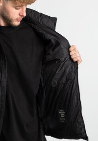 Zwarte geïsoleerde jas met een rits, gekenmerkt door een geborduurde buitenkant en een gladde binnenkant. Heeft een label met de tekst "Koude Weer Warm."