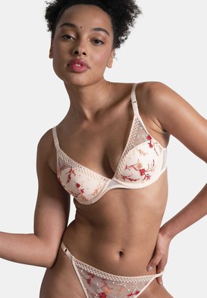Femme portant une lingerie beige transparente avec une broderie florale rouge et orange, posant avec une main sur la hanche sur un fond uni.