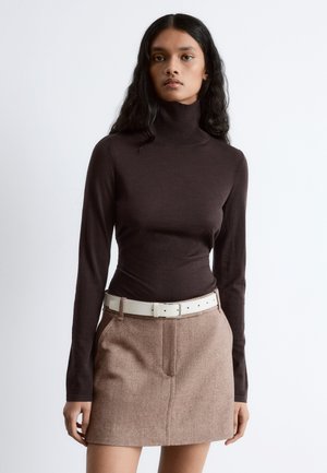 Brauner Rollkragenpullover in taillierter Passform, kombiniert mit einem beigen, gewebten Rock, der zwei Taschen und einen weißen Gürtel hat. Glatte Textur, moderner Look.