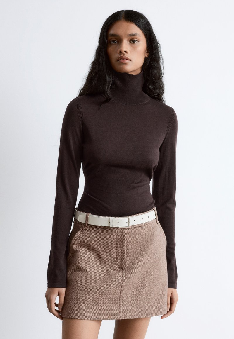 Brown turtleneck sweater in a fitted design paired with a beige woven skirt featuring two pockets and a white belt. Smooth texture, modern look.