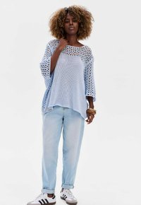 Jasnoniebieski sweter dzianinowy o luźnym, otwartym fasonie i szerokich rękawach, zestawiony z jasnoniebieskimi dżinsami i białymi sneakersami w czarne paski.