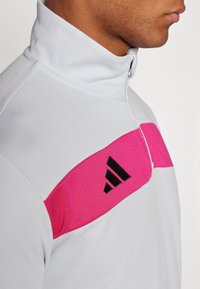 Veste de sport à col haut gris clair avec un panneau rose horizontal et un logo noir. Texture lisse avec une fermeture zippée.