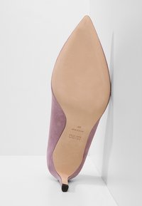 Chaussure à talon haut en daim mauve à bout pointu présentée semelle vers le haut, montrant la semelle en cuir et la petite pointe de talon noire.