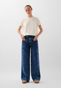 Hellblaue, hochtaillierte Jeans mit weitem Bein, kombiniert mit einem cremefarbenen T-Shirt mit kurzen Ärmeln. Die Jeans haben ein einzigartiges Nahtdesign und sichtbare Stiche.