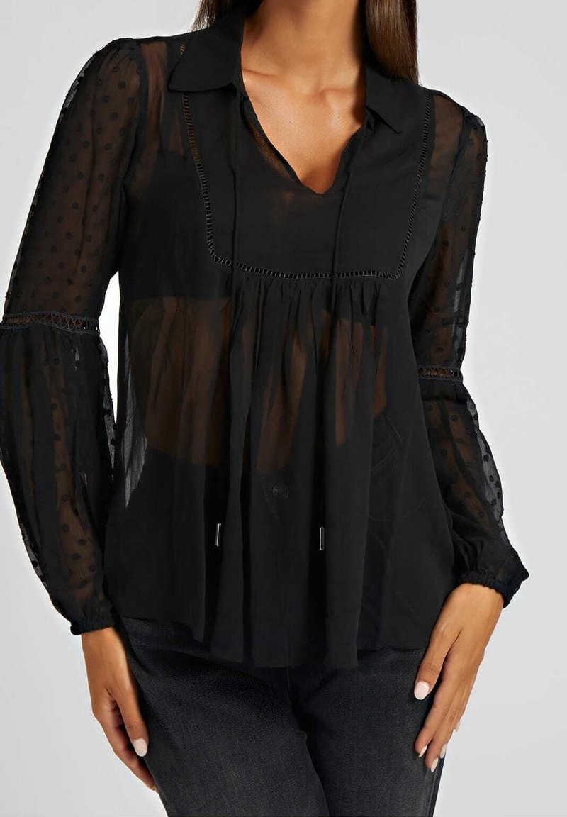 Blouse noire transparente avec col, texture à pois, plis couchés et manches froncées. Présente une coupe décontractée et des fermetures à boutons discrètes.