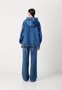 Denim oversized jacka med huva, blå färg, avslappnad passform och vida denimbyxor. Synliga sömdetaljer och uppvikt byxslut.