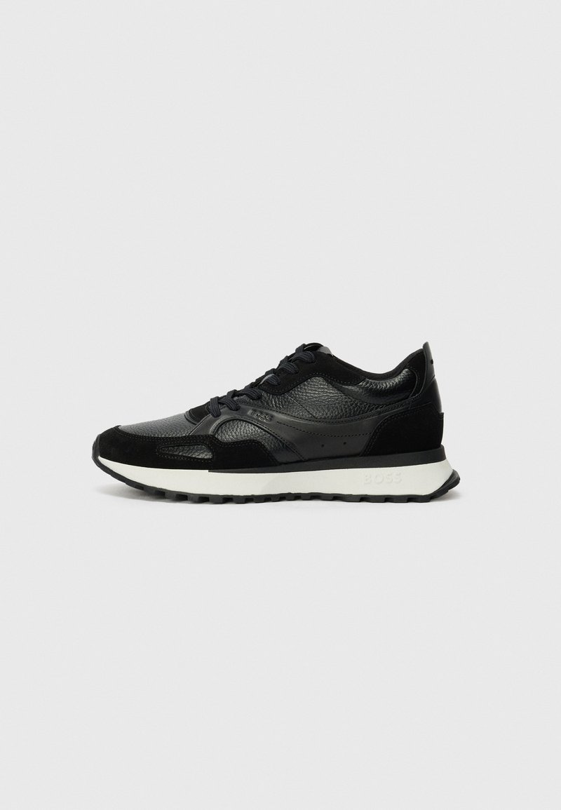 Baskets noires en cuir texturé et en daim, avec une semelle intermédiaire blanche et un design à lacets. Logo de marque notable sur le côté.