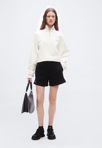 Γυναίκα με λευκή φούτερ με φερμουάρ και μαύρο σορτς, κρατώντας μαύρη τσάντα, στέκεται μπροστά σε απλό λευκό φόντο.