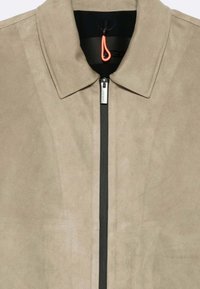 Giacca in suede beige con zip frontale nera, colletto a punta e dettagli arancioni a forma di cerchio sul collo. Texture liscia, design minimalista.