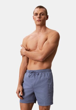 Giovane uomo con capelli corti che indossa pantaloncini da bagno blu Calvin Klein, in piedi a torso nudo con un braccio incrociato sul petto.