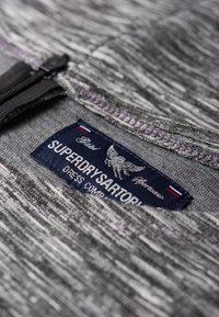 Šedá texturovaná tkanina se zipem a námořnickou etiketou s nápisem "SUPERDRY SARTORIA", která obsahuje logo orla a červené a bílé akcenty.