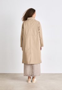 Beige, knie-length jas met een hoge kraag, getailleerde taille en gestikte details. Gecombineerd met een lichtgrijze denim rok en casual schoenen.