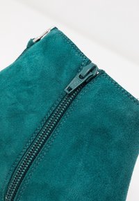 Sinisenvihreä mokkanahkainen pussukka, jossa näkyvä vetoketjun yksityiskohta. Sileä pinta, suorakaiteen muotoinen, vankka ompelu ja korostettu vetoketjun vedin.