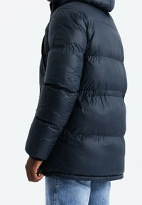 Giacca a vento imbottita blu navy con cappuccio, caratterizzata da una texture liscia e lucida, pannelli cuciti e una piccola patch con logo sulla manica sinistra.