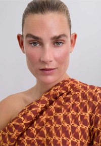 Wikkel in bruin met een oranje geometrisch patroon, met een gestructureerde stof die over één schouder valt. Soepele huidtinten en natuurlijke make-up.