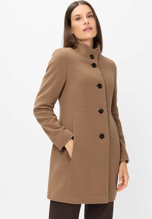 Woman wearing a knee-length brown coat with high collar and five black buttons, one hand in pocket, standing against plain light background.