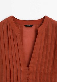 Rust-orange bluse med lodrette folder, en rund halsudskæring, og fem knapper foran. Fremstillet af letvægtsstof.