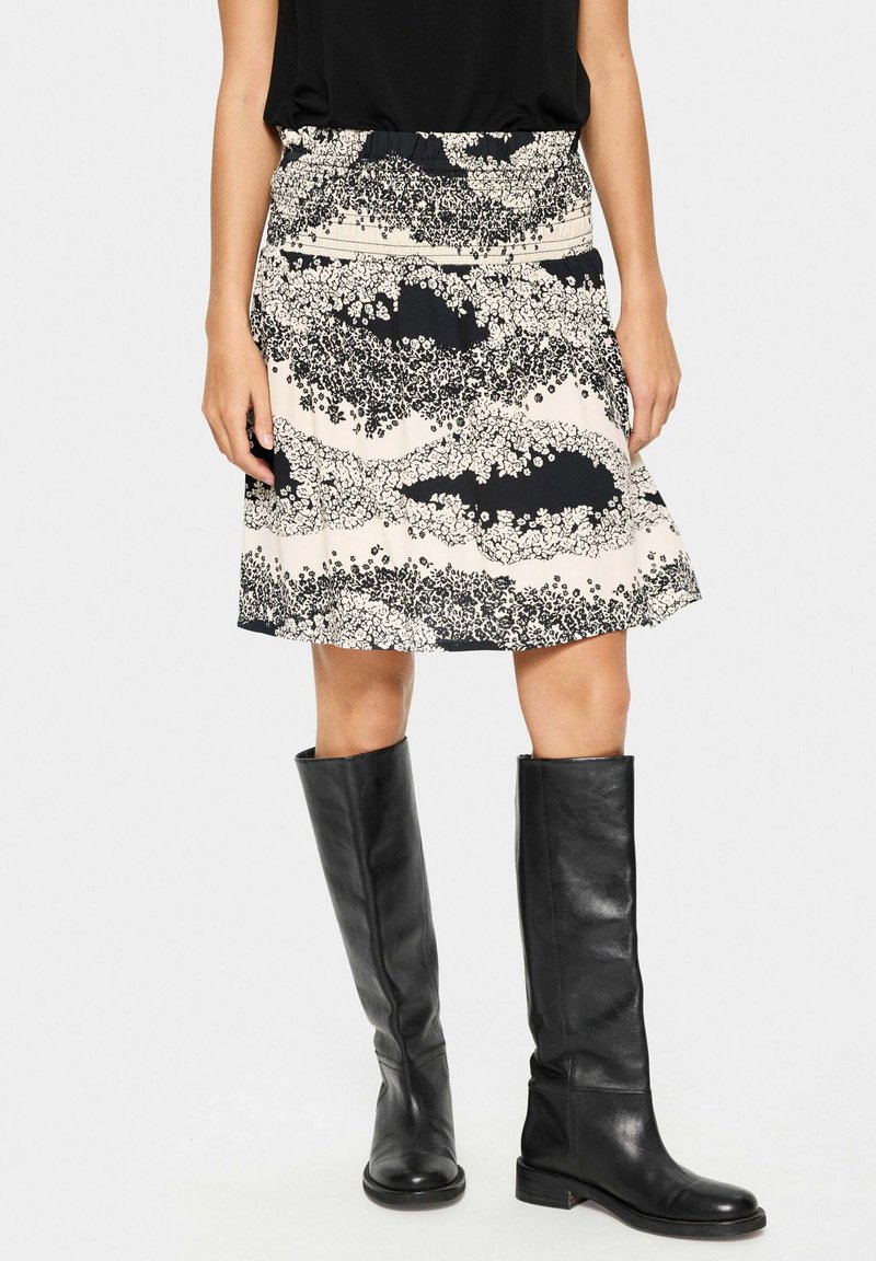 Jupe à motif floral en crème et noir, fabriquée en tissu léger avec une taille élastique, associée à des bottes en cuir noires jusqu'aux genoux.