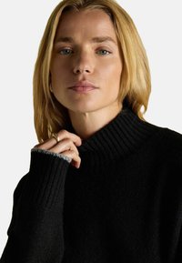 Melnā augstās apkakles džemperis ar ribotām tekstūrām un pelēkiem akcentiem pie aprocēm. Modelis viegli tur apkakles daļu ar vienu roku. Gluda auduma apdare.