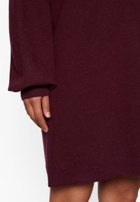 Burgunderfarbiges Strickkleid mit langen, lockeren Ärmeln. Der Stoff wirkt weich, hat eine glatte Textur und einen leicht entspannten Schnitt, der oberhalb des Knies endet.