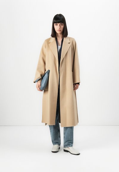 Kvinna med rakt mörkt hår iklädd en lång beige kappa, blå jeans, vita skor och håller en blå handväska framför en neutral vit bakgrund.