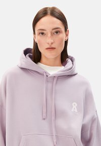 Lavender Kapuzenpullover aus weichem Stoff, mit einer Kängurutasche und einem kleinen weißen Logo auf der linken Brust. Kordelzüge an der Kapuze.