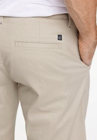 Pantaloni beige a trama con un motivo geometrico, dotati di tasca posteriore e di una piccola etichetta nera vicino alla vita, tenuti in mano.