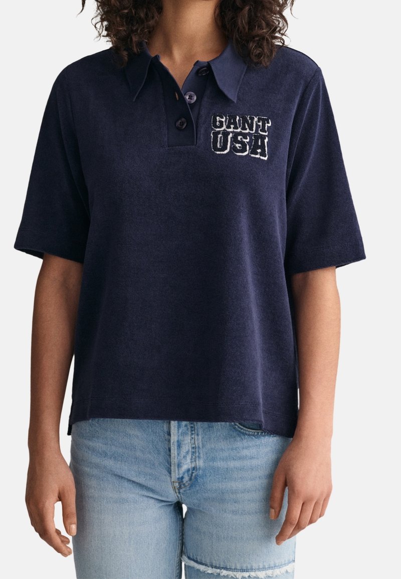 Personne portant un polo bleu marine à manches courtes avec le logo "GANT USA" sur le côté gauche de la poitrine et un jean bleu clair déchiré.