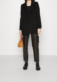 Blazer di lana nera con rever a punta, abbinato a pantaloni in pelle nera lucida. Gli accessori includono una piccola borsa gialla e stivali neri massicci.