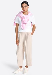 Tricou alb cu un design imprimat, hanorac roz deschis purtat pe umeri, pantaloni bej plisati și adidași albi cu accente roz.