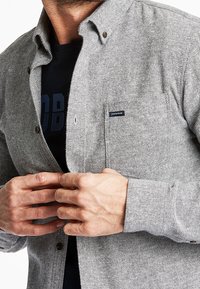 Camicia a spina di pesce grigia con colletto button-down, tasca frontale e bottoni marroni. Indossata sopra una t-shirt nera con grafica.