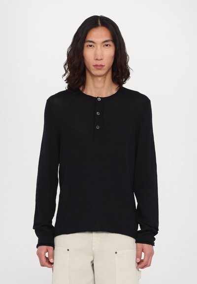 Chemise noire en maille à manches longues avec un col rond, une patte de boutonnage à trois boutons et une coupe décontractée, associée à un pantalon cargo de couleur claire.
