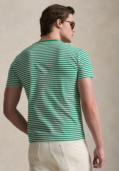 Mees, kes kannab rohevalge triibulist lühikeste varrukatega särk ja valgeid pükse, näoga ära, päikeseprillid peas ja lühikeste pruunide juustega.