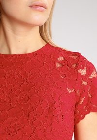 Červený krajkový top s květinovými vzory, krátkými rukávy a kulatým výstřihem, prezentující texturovaný, propracovaný design a přiléhavý střih.