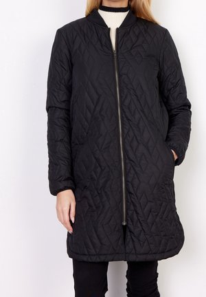 Femme portant une longue doudoune noire avec une fermeture éclair devant et des poches, associée à un pantalon noir et un pull blanc à col montant.
