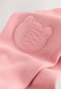 Material dintr-un hanorac roz, cu o patch rotund și pufos în formă de față de urs, contururi cusute, prezentând o textură moale și un finisaj neted.