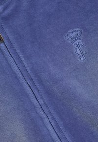 Tissu polaire bleu avec une fermeture éclair visible et un logo brodé montrant une couronne au-dessus des lettres stylisées "J" et "C".