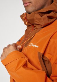 Orange wasserfeste Jacke mit braunen Akzenten, ausgestattet mit einem hohen Kragen, verstellbaren Kordelzügen und einem sichtbaren Logo auf der Brust.