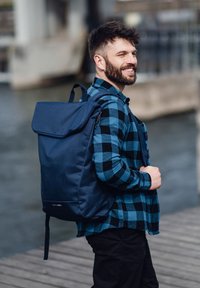 Marinefarbene Rucksack aus robustem Stoff, mit einem umklappbaren Deckel, verstellbaren Trägern und einem Logo auf der Vorderseite. Getragen von einer lächelnden Person.