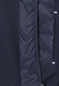 Mornarska jakna koja kombinira glatku tkaninu i kviltanu teksturu, s punim patentnim zatvaračem i suptilnim akcentima kamuflažnog uzorka unutar jakne.