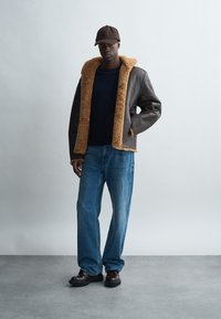 Brown leather jacket with tan faux fur collar, blue jeans, and dark shoes. Model stands against a light gray background, wearing a dark cap.