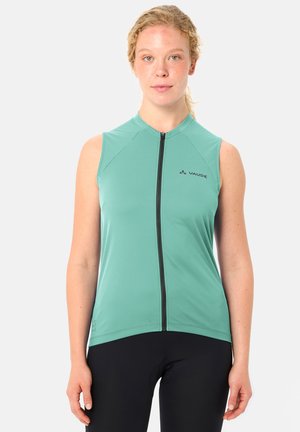 Femme aux cheveux blonds bouclés portant un haut de sport turquoise sans manches avec fermeture éclair et des leggings noirs, debout devant un fond neutre.