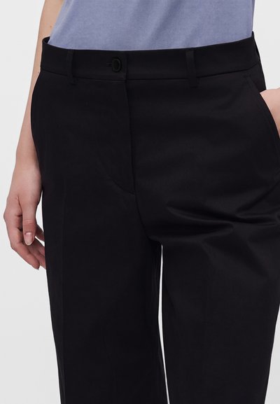 Personne portant un pantalon noir sur mesure et un haut bleu clair, debout avec une main dans la poche et l'autre détendue sur le côté.