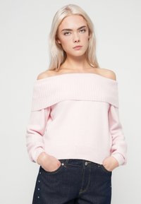 Ανοιχτό ροζ ελκυστικό πουλόβερ με εκτεθειμένους ώμους, ριμπ κόψιμο στον λαιμό, μακριά μανίκια και μαλακή υφή, σε συνδυασμό με σκούρες τζιν παντελόνες.