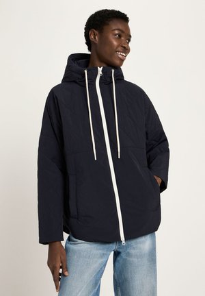 Lächende Person, die eine schwarze Kapuzenjacke mit Reißverschluss und weißen Kordeln sowie hellblaue Jeans trägt, vor einem einfarbigen Hintergrund stehend.