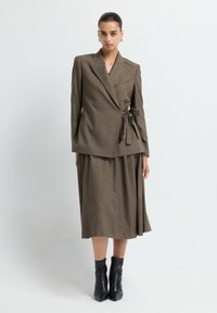 Traje de dos piezas a rayas marrones con una chaqueta cruzada que cuenta con un lazo en la cintura y una falda fluida, combinado con botas negras hasta el tobillo.