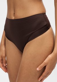 Femme portant un bas de bikini taille haute marron foncé, visible de la taille au milieu de la cuisse sur un fond uni.