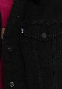 Czarna dżinsowa kurtka z guzikowym zapięciem z przodu, dwoma kieszeniami na klatce piersiowej i miękkim, teksturowanym wnętrzem. Minimalistyczny design bez wyraźnych wzorów.